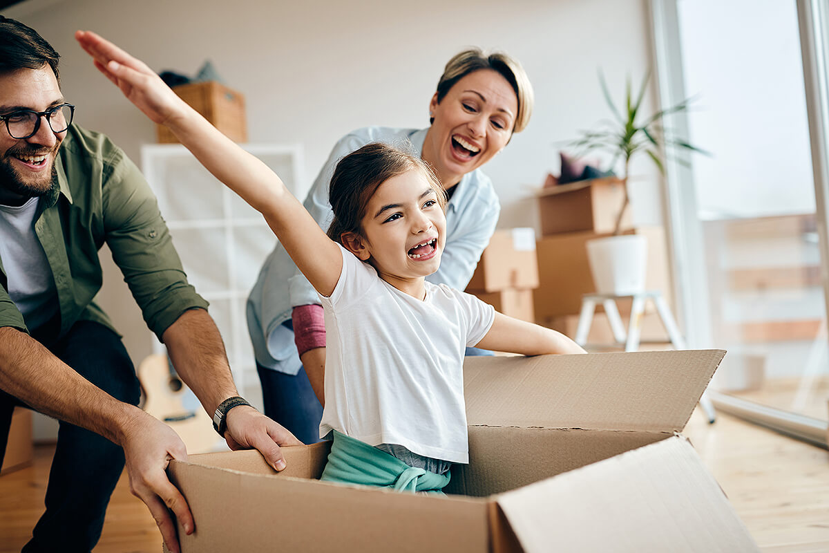 Young girl in box with arms out while mum and dad push her around laughing