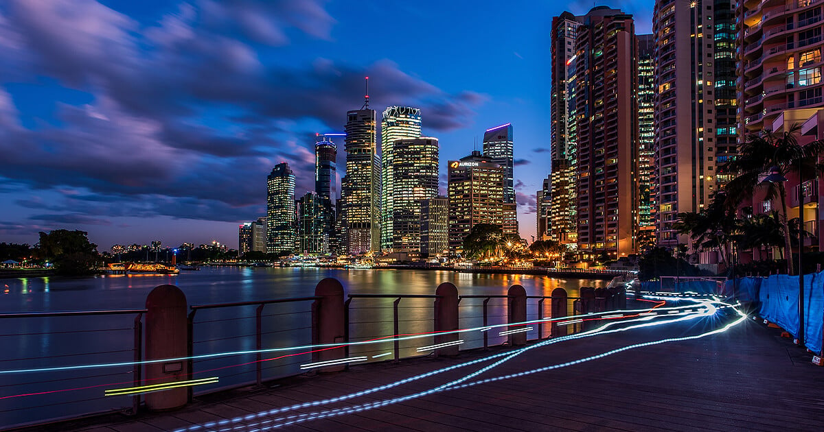 Brisbane city skyscrapers reflecting on river and bike light trails on sidewalk at dusk