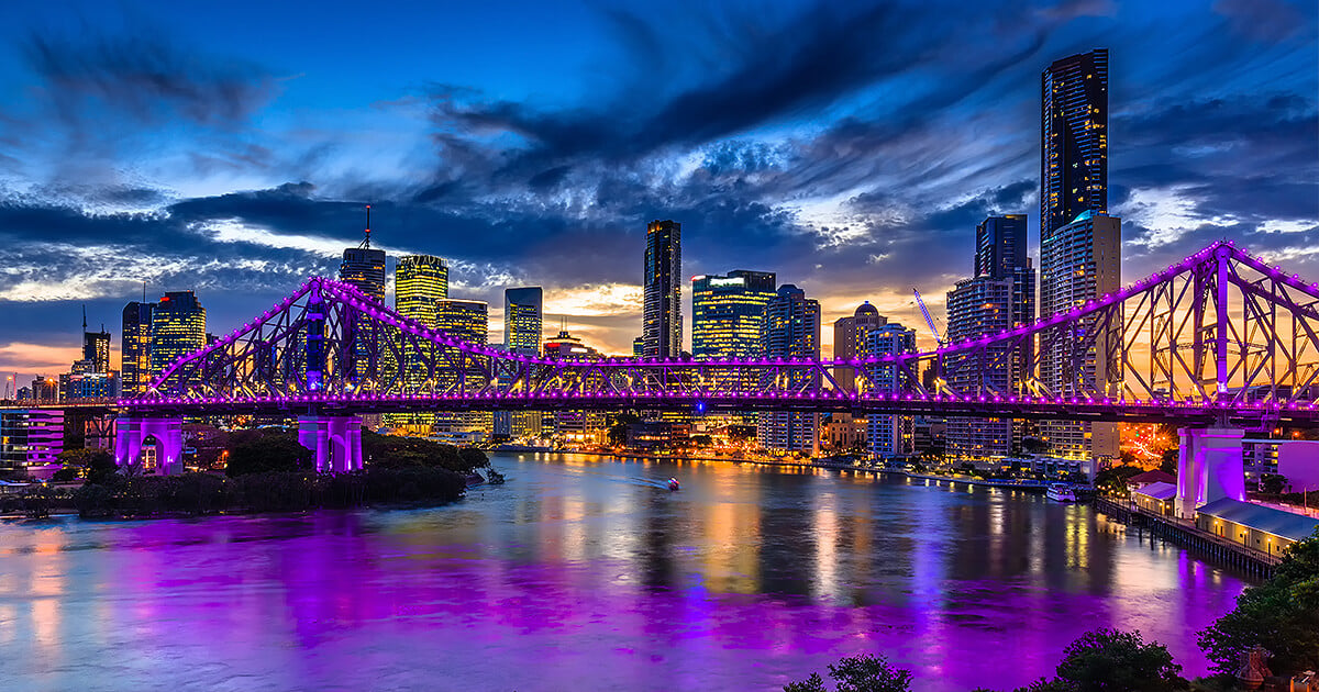 The Story Bridge lit up in purple with Brisbane city in the background at dusk