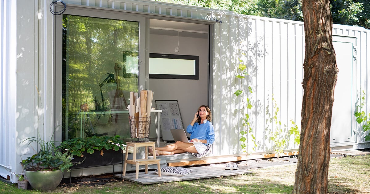 Woman sits in doorway of shipping container home office while on laptop doing work