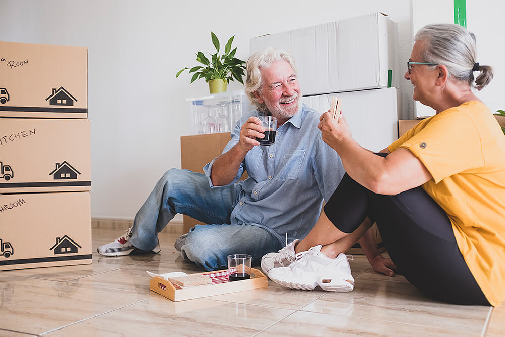 Happy couple drinking wine and eating while sitting on the floor surrounded by boxes after moving to Brisbane